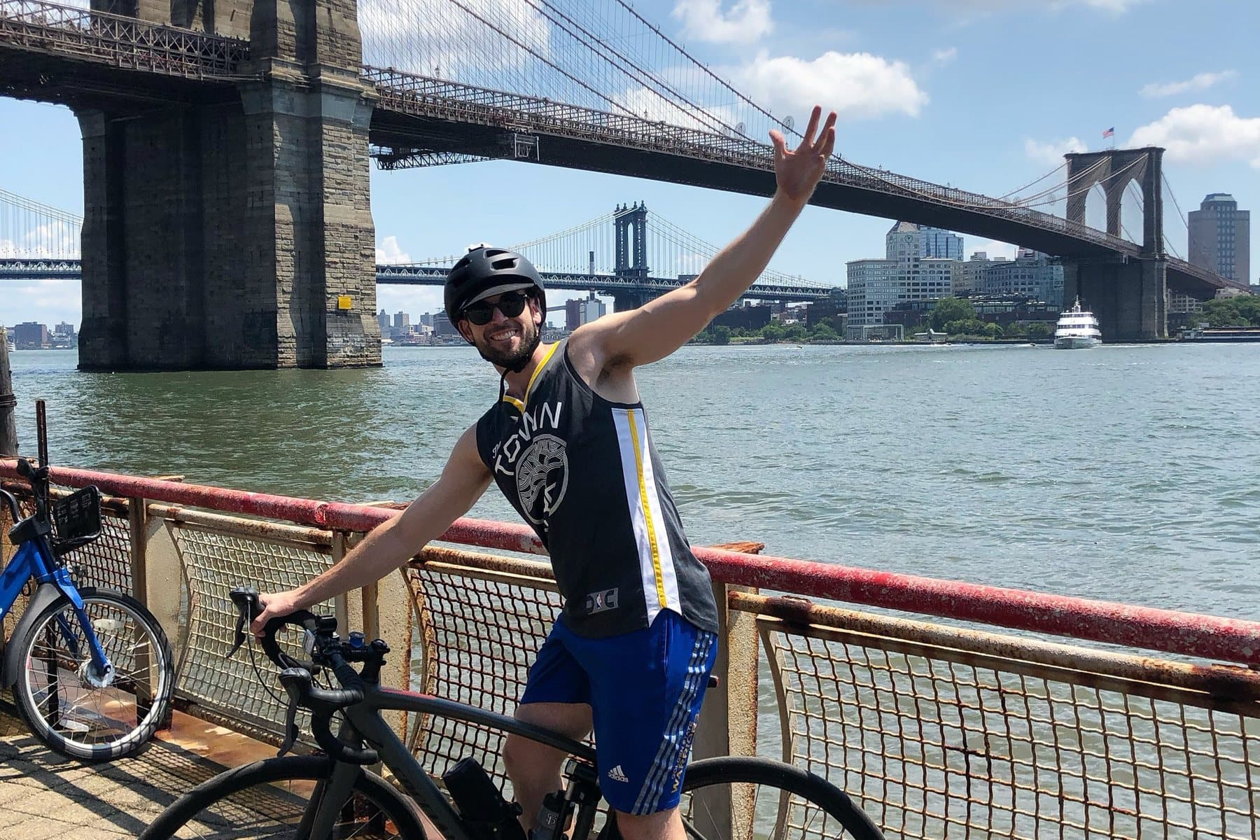 Daniel waving from his bike on the East River waterfront, with the Brooklyn and Manhattan Bridges behind him